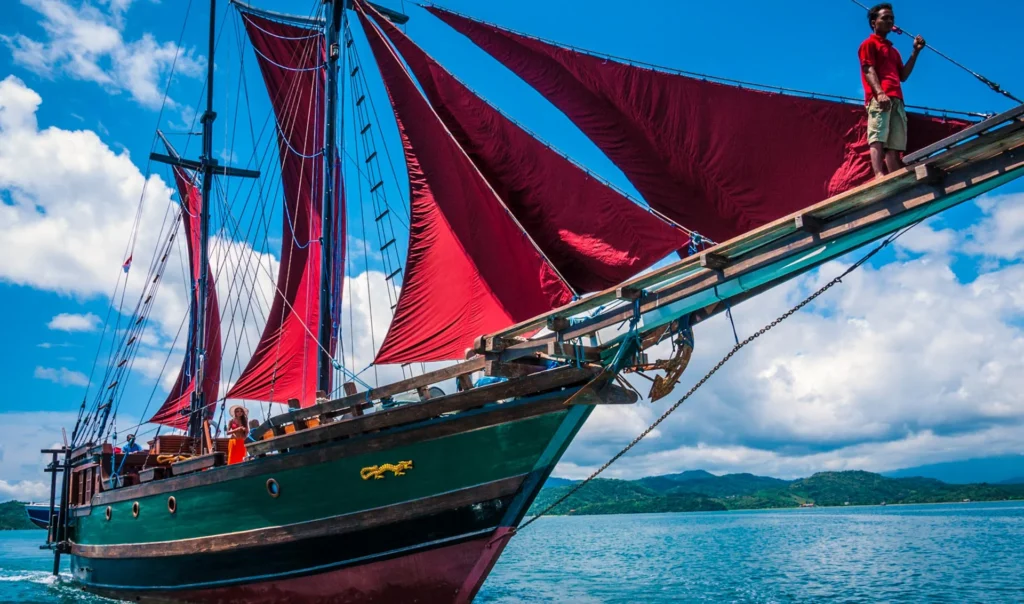 Ananda phinisi under red sails with a crew member standing on the bowsprit