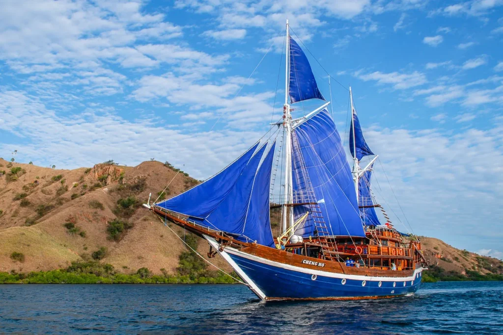 Cheng Ho phinisi with blue sails raised passing dry, grassy hills