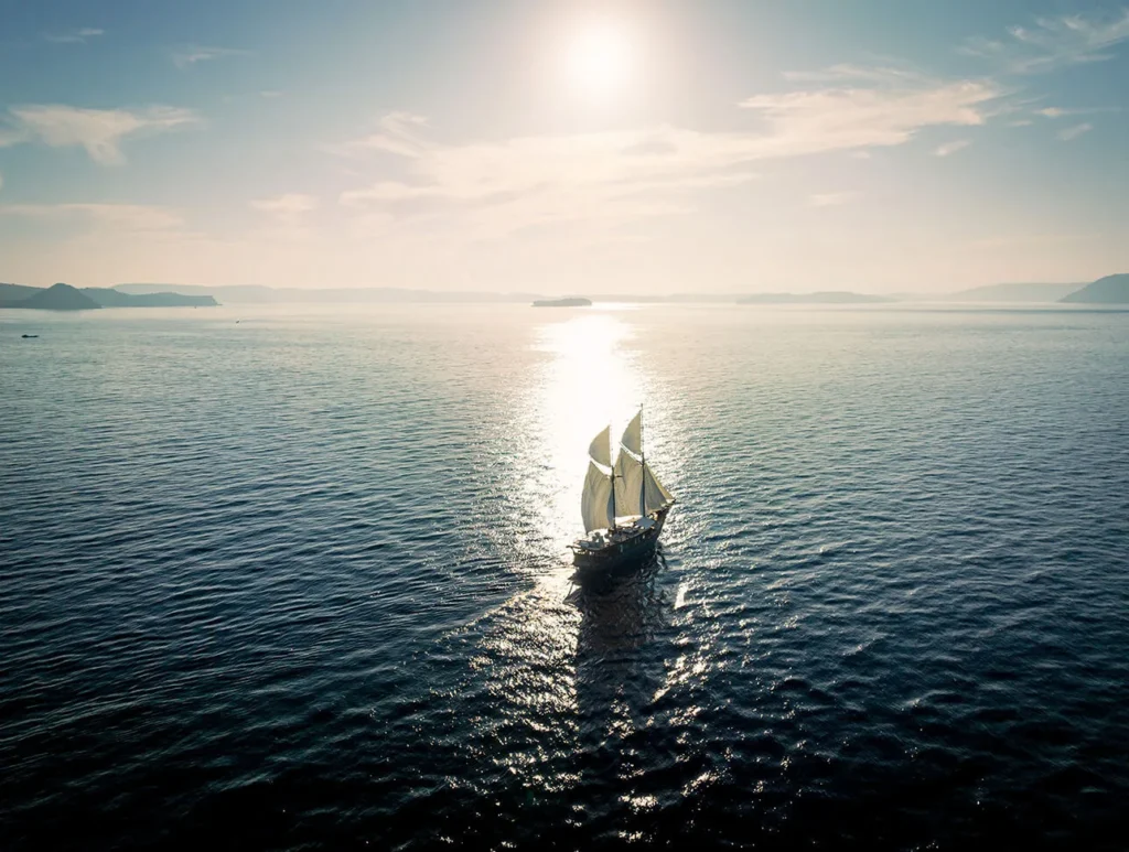 Dewata under sail on open water with sunlight glinting on the sea