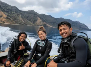 Three smiling scuba divers wearing wetsuits sitting on a moving speedboat against the backdrop of dry hills in Komodo National Park.