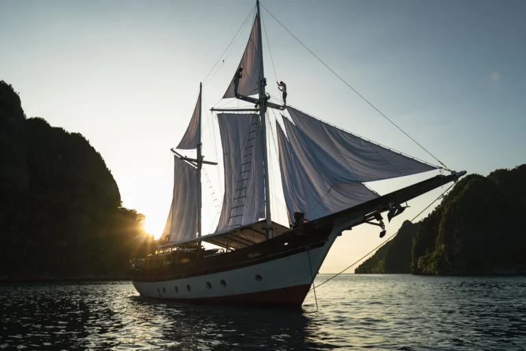 Anne Bonny phinisi under white sails between rocky islands at sunset
