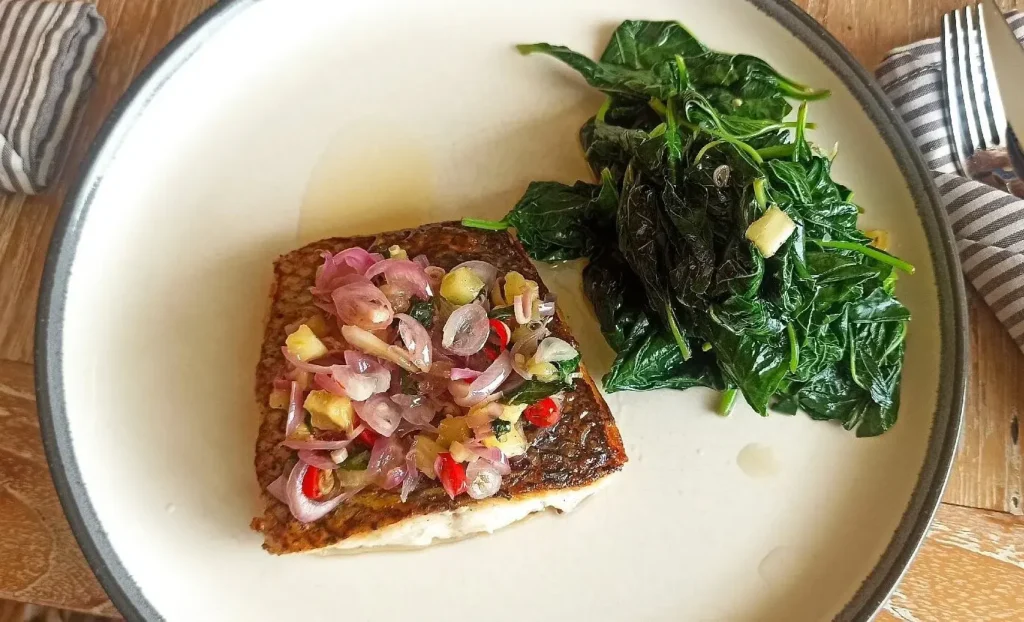 Seared fish fillet topped with salsa and served with sauteed greens on a white plate