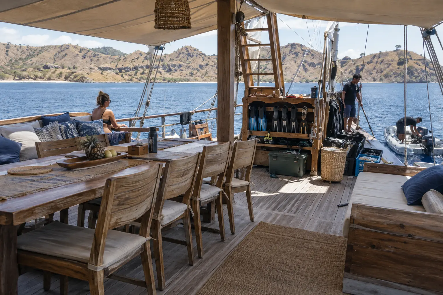 A phinisi deck with dining space, shaded lounge, snorkel gear and a tender being prepared.