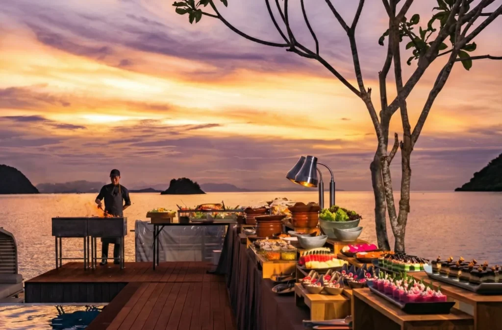 Buffet dishes laid out on a deck at sunset with islands behind at Layar Restaurant